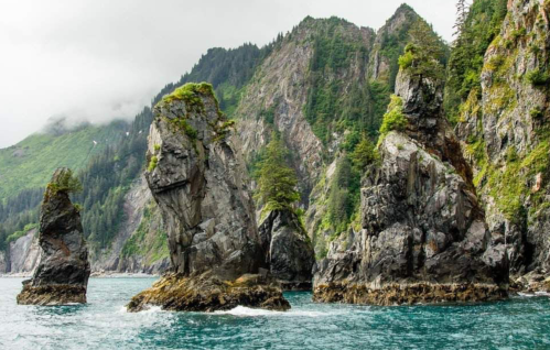 Rocky coastal formations rise from calm waters, surrounded by lush green hills and a cloudy sky.