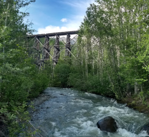 A rushing river flows through a lush green landscape, with a tall wooden trestle bridge in the background.