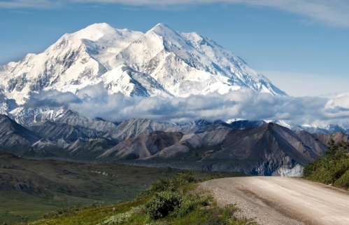 A scenic view of a snow-capped mountain range with a gravel road in the foreground and blue skies above.