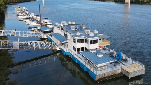Aerial view of a floating restaurant on a river, surrounded by boats and a dock, with a blue roof and outdoor seating.