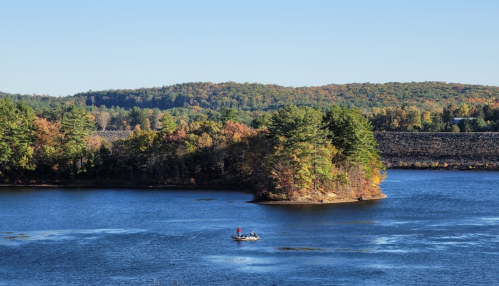 A serene lake scene with a small boat and an island, surrounded by trees in autumn colors under a clear blue sky.