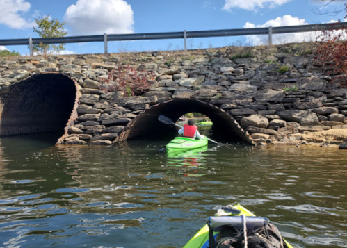 A person in a green kayak paddles through a stone archway under a bridge on a calm waterway.