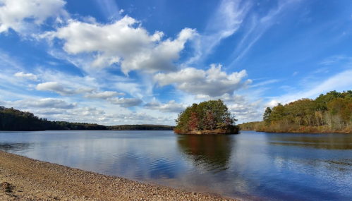 A serene lake scene with a small island, surrounded by trees and a blue sky filled with fluffy clouds.
