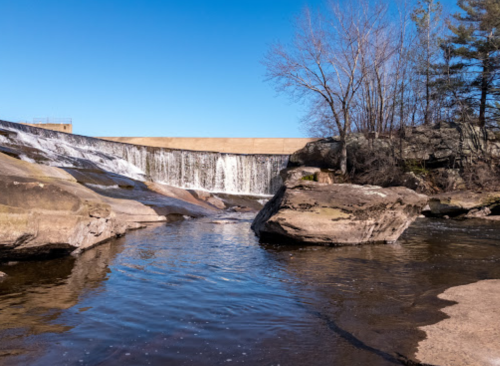 A serene waterfall cascades over rocks, surrounded by trees under a clear blue sky.