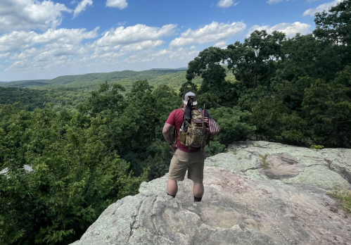A hiker stands on a rocky ledge, overlooking a lush green valley under a partly cloudy sky.