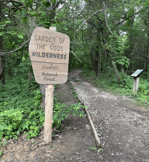 Wooden sign for "Garden of the Gods Wilderness" at Shawnee National Forest, with a gravel path surrounded by greenery.