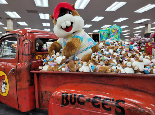 A large mascot in a colorful shirt sits in a vintage red truck filled with plush toys, inside a store.