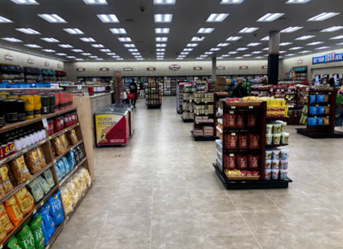 A spacious grocery store aisle with shelves stocked with snacks, beverages, and various food items under bright lights.