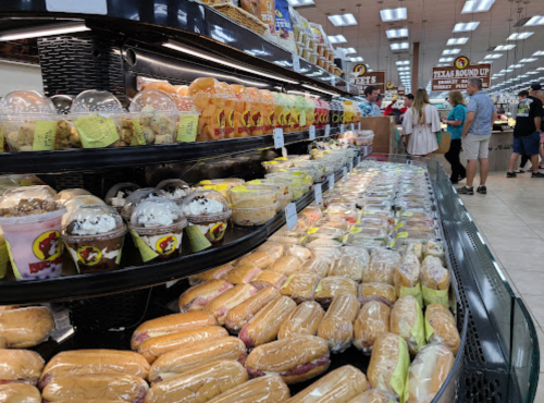 A display of various hot dogs and desserts in a grocery store, with shoppers in the background.