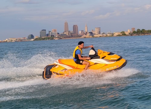 A person rides a yellow jet ski on water with a city skyline in the background on a sunny day.