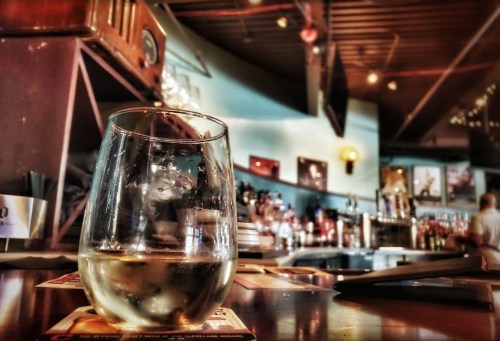 A close-up of a glass of white wine on a bar counter, with a blurred background of a cozy bar setting.
