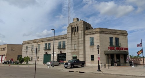 Historic fire museum building with a tall tower, large windows, and flags outside, set against a cloudy sky.