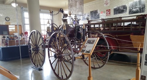 A vintage fire engine on display in a museum, featuring wooden wheels and brass fittings, with historical photos in the background.