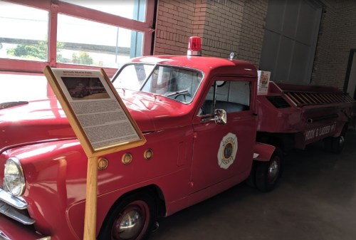 A vintage red fire truck with a light on top, displayed indoors next to an informational sign.