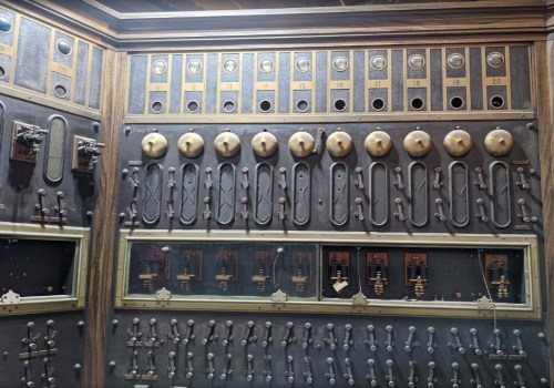 A vintage control panel with numerous switches, knobs, and labeled buttons arranged in rows on a dark background.
