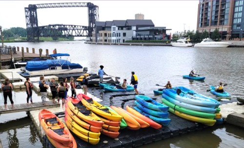 A group of people preparing to kayak near a dock with colorful kayaks and a bridge in the background.