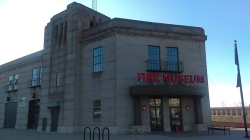 Exterior of a stone building with large windows, featuring a sign that reads "FIRE MUSEUM" in bold red letters.