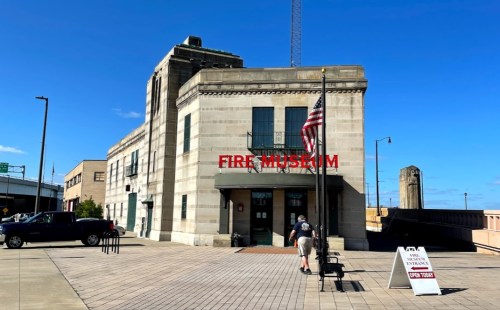 Exterior of a fire museum with a flag, sign reading "FIRE MUSEUM," and a visitor approaching the entrance.