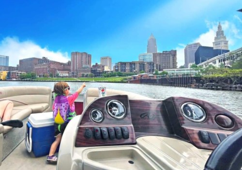 A child in a colorful shirt sits on a boat, looking at a city skyline under a bright blue sky.
