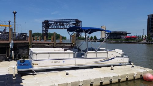 A pontoon boat docked at a marina with a blue canopy, surrounded by calm water and a bridge in the background.