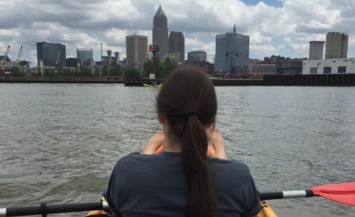 A person in a kayak faces a city skyline with tall buildings under a cloudy sky.