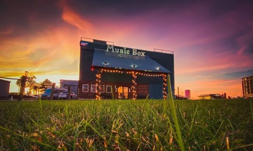 A vibrant sunset behind a building labeled "Music Box," with string lights and green grass in the foreground.