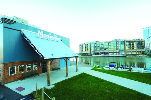 A riverside view of the Music Box building with modern apartments in the background and a grassy area in the foreground.
