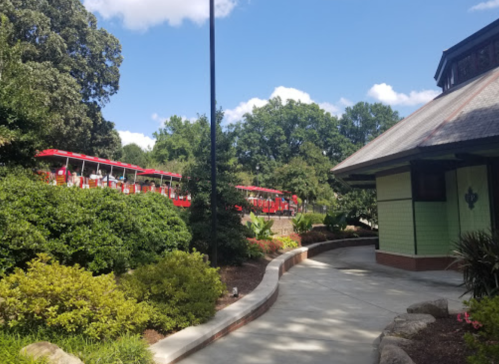 A winding path leads to a green building, with red trams visible in the background among lush trees and blue skies.