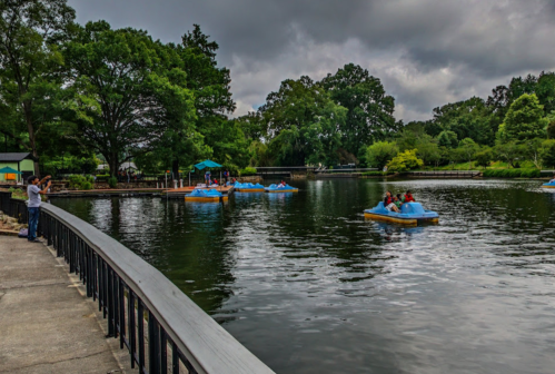 People enjoy paddle boating on a serene lake surrounded by lush greenery under a cloudy sky.
