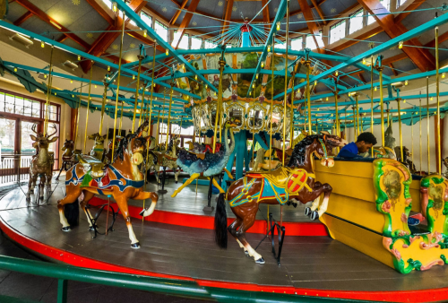A colorful carousel with ornate horses and a central chandelier, set in a bright, spacious pavilion.