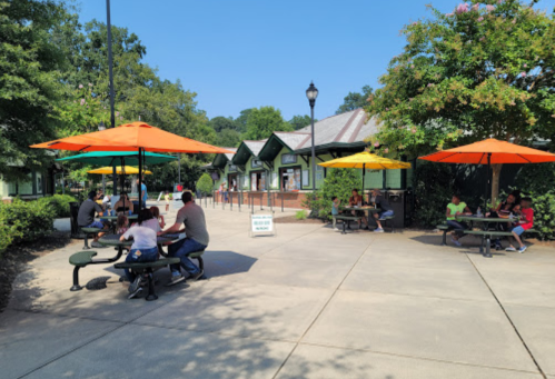Outdoor dining area with colorful umbrellas, people sitting at tables, and green trees in a sunny park setting.