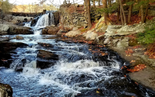 A serene waterfall cascades over rocks, surrounded by autumn foliage and a tranquil stream.