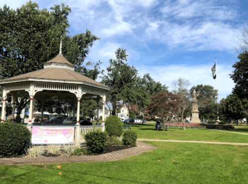A gazebo in a park with trees, a flagpole, and a monument, under a blue sky with scattered clouds.