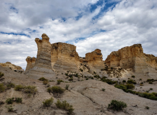 Rocky formations rise against a cloudy sky, surrounded by sparse vegetation in a desert landscape.
