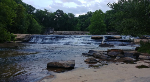 A serene river scene with gentle waterfalls, surrounded by lush greenery and rocky banks under a cloudy sky.