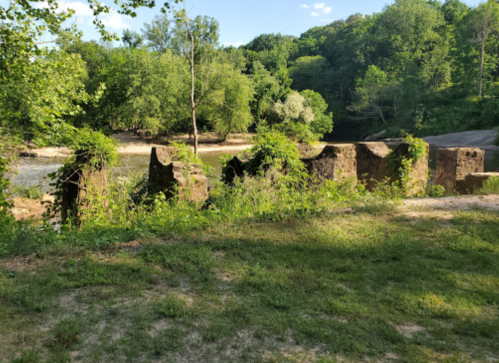 Overgrown stone ruins by a river, surrounded by lush greenery and trees under a clear blue sky.