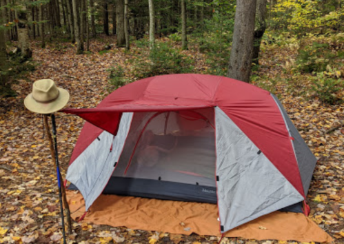 A red and gray tent set up in a forest with fallen leaves, accompanied by a hat and walking stick nearby.