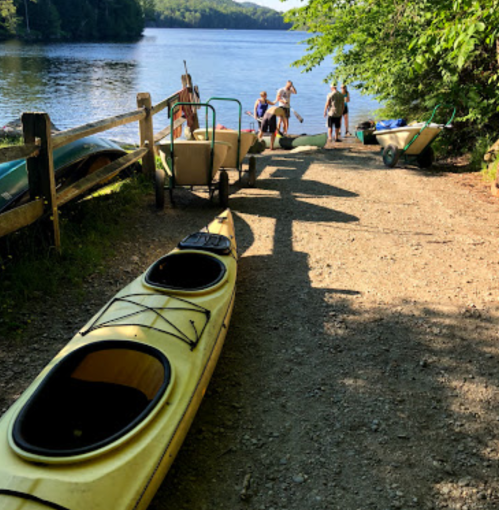 A sunny lakeside scene with kayaks on the shore and people preparing to launch them into the water.