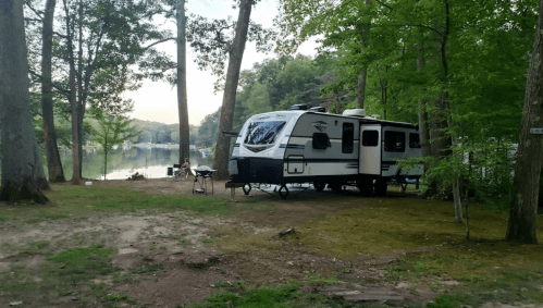 A camper parked by a serene lake, surrounded by trees, with a person sitting nearby.
