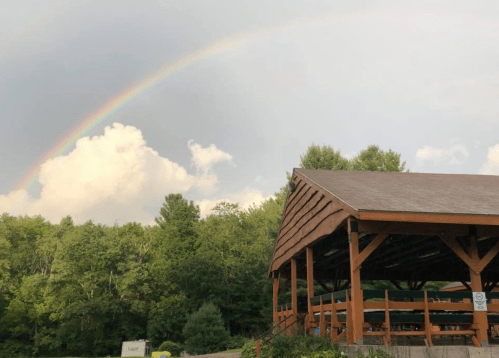 A wooden pavilion under a rainbow with lush green trees in the background and a cloudy sky.