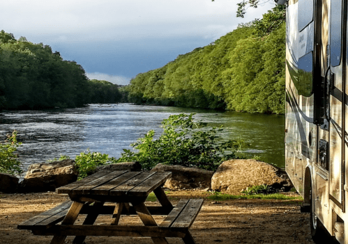 A serene river scene with lush greenery, a picnic table, and an RV parked nearby under a cloudy sky.