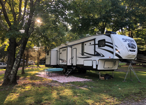 A white RV parked under trees, with a picnic area set up on the grass beside it. Sunlight filters through the leaves.
