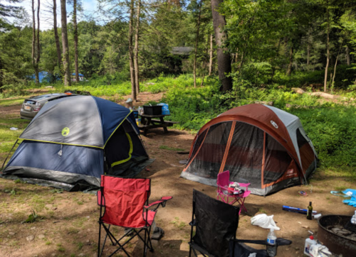 Two tents set up in a wooded campsite, with camping chairs and a picnic table nearby.