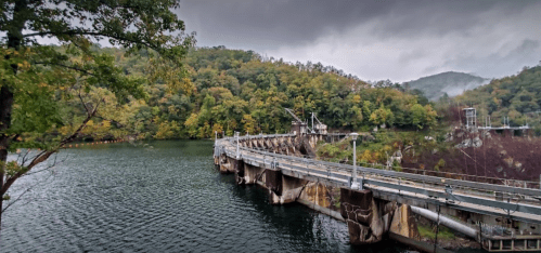 A panoramic view of a dam surrounded by lush green hills and calm water under a cloudy sky.