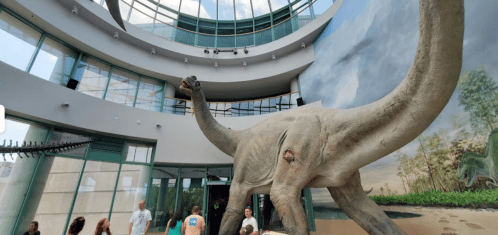 A large dinosaur model stands in a museum atrium, with visitors admiring it and a mural of nature in the background.