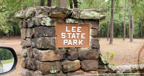 Sign for Lee State Park on a stone structure, surrounded by trees and a gravel area.