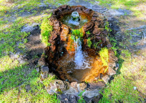 A small stone fountain with water flowing over rocks, surrounded by green grass in a sunny outdoor setting.