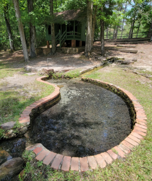 A curved brick-lined stream flows through a grassy area, with a wooden cabin visible in the background among trees.