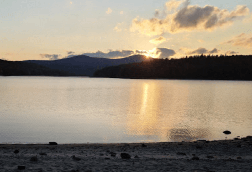 Sunset over a calm lake, with mountains in the background and soft clouds in the sky, reflecting on the water's surface.