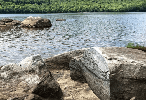 A serene lakeside scene featuring large rocks and calm water, surrounded by lush green trees in the background.
